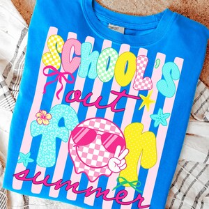 May include: A blue t-shirt with the words "School's Out For Summer" in a colorful, playful font. The design includes pink stripes, stars, and a smiley face wearing sunglasses. The shirt is a comfortable, casual style.