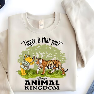 Puede incluir: Una sudadera beige con un gr&aacute;fico de Winnie de Pooh mirando a un tigre con una lupa. El texto "Tigger, is that you?" est&aacute; por encima del gr&aacute;fico. Debajo del gr&aacute;fico est&aacute; el texto "Disney's Animal Kingdom".
