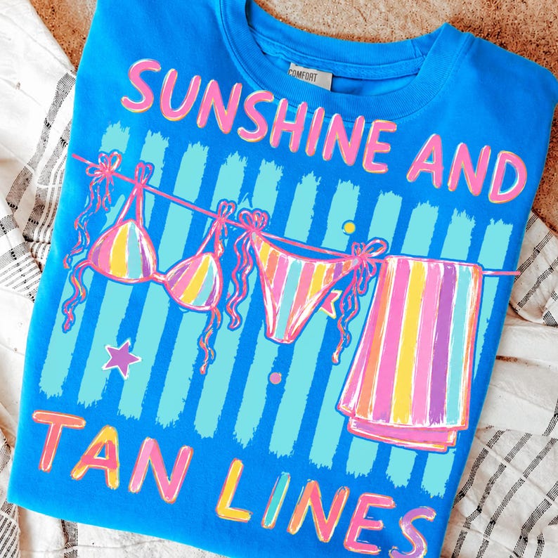 Peut inclure: Un t-shirt bleu avec les mots "SUNSHINE AND TAN LINES" en rose et jaune. Le t-shirt pr&eacute;sente un motif de bikini et de serviette suspendus &agrave; une corde &agrave; linge, avec des rayures color&eacute;es et une &eacute;toile.