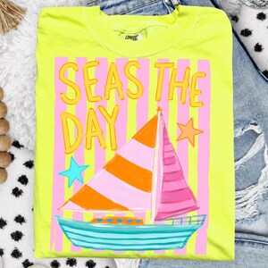 May include: A neon yellow t-shirt with the phrase "SEAS THE DAY" in yellow bubble letters. The shirt features a sailboat with orange and pink sails, a turquoise hull, and pink and yellow striped background.