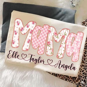 May include: Cream-colored t-shirt with the word "Mama" in large, decorative letters. The letters feature floral, checkered, and striped patterns in pink and white. Below the word are the names "Elle", "Taylor", and "Angela" in cursive script.