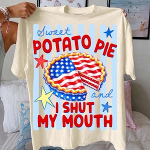 May include: Cream-colored t-shirt featuring a graphic of a sweet potato pie with red, white, and blue accents. The text on the shirt reads "Sweet Potato Pie and I Shut My Mouth" with star accents. The shirt is held up by a person.