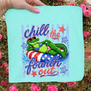 May include: A light blue square towel with a graphic of an alligator wearing sunglasses and a bikini, lounging on a red, white, and blue inner tube. The text "Chill the Fourth Out" is written above and below the alligator.