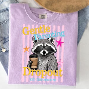 Op de afbeelding: Een lila Comfort Colors t-shirt met een wasbeer met een zonnebril en een koffiekopje. Het shirt heeft de woorden "Gentle Parenting Dropout" en "Est. This Morning" in een kleurrijk ontwerp met sterren.
