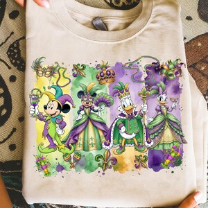 May include: Beige t-shirt featuring a colorful graphic of Disney characters dressed in Mardi Gras attire. Mickey Mouse, Minnie Mouse, Donald Duck, and Daisy Duck are depicted in festive costumes with masks, crowns, and fleur-de-lis symbols.