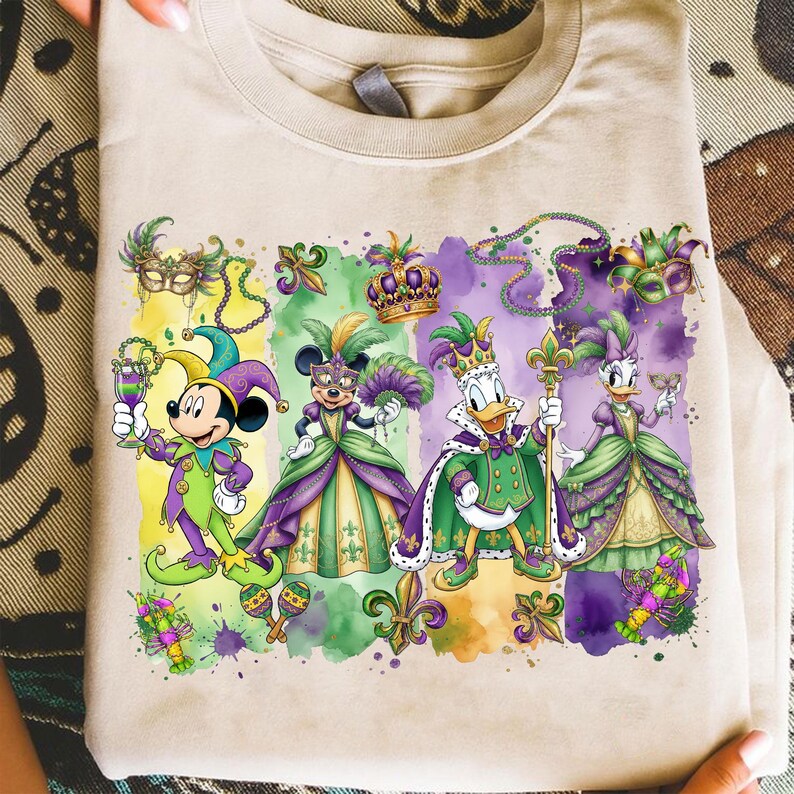 May include: Beige t-shirt featuring a colorful graphic of Disney characters dressed in Mardi Gras attire. Mickey Mouse, Minnie Mouse, Donald Duck, and Daisy Duck are depicted in festive costumes with masks, crowns, and fleur-de-lis symbols.