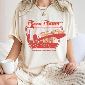 Puede incluir: Camiseta color crema con un gr&aacute;fico retro de Pizza Planet en rojo y amarillo. El dise&ntilde;o incluye un cohete, un restaurante y el texto "Pizza Planet" y "Serving Your Local Star Cluster."