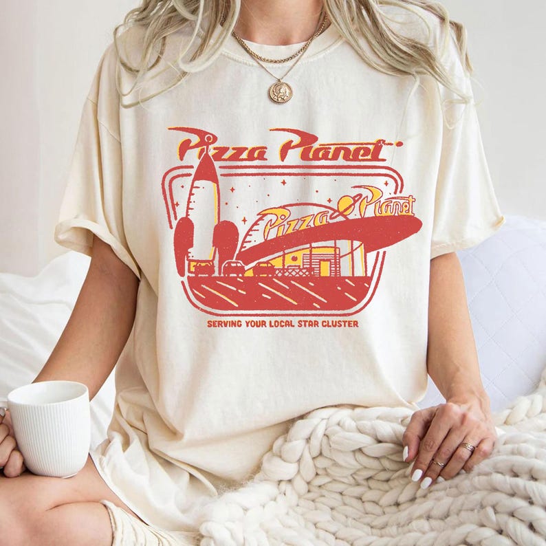 Puede incluir: Camiseta color crema con un gr&aacute;fico retro de Pizza Planet en rojo y amarillo. El dise&ntilde;o incluye un cohete, un restaurante y el texto "Pizza Planet" y "Serving Your Local Star Cluster."