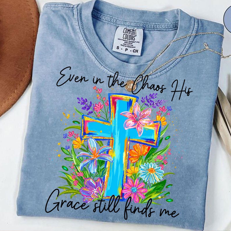May include: A light blue t-shirt with the text "Even in the Chaos His Grace still finds me." The shirt features a colorful cross design surrounded by flowers in shades of pink, blue, yellow, and purple. A gold necklace is draped across the shirt.