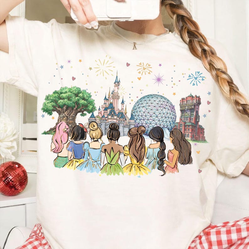 May include: Cream-colored t-shirt featuring a colorful illustration of several cartoon princesses with their backs to the viewer, in front of iconic theme park landmarks. The design includes a castle, a large sphere, and a tree.