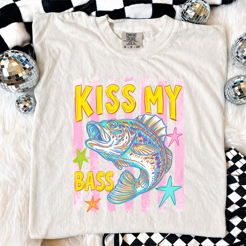 May include: White t-shirt with the text "KISS MY BASS" in yellow letters above a colorful fish illustration. Pink vertical stripes and star graphics surround the fish. The shirt is a Comfort Colors brand.