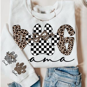May include: White sweatshirt with the words "Dog Mama" in a mix of leopard print and checkered paw prints. A smaller sweatshirt shows paw prints with names. The design includes a bow and cursive font.