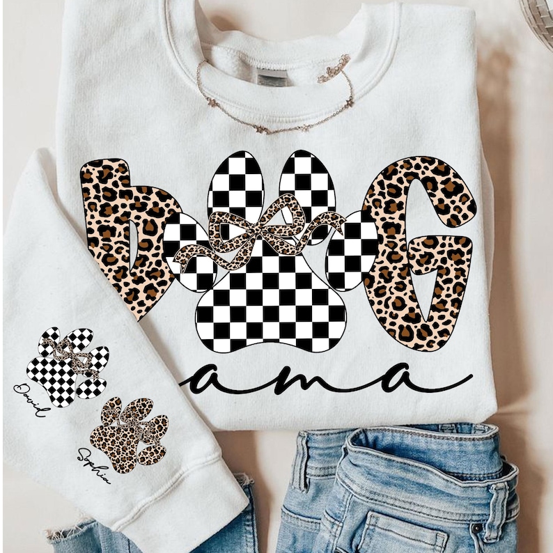 May include: White sweatshirt with the words "Dog Mama" in a mix of leopard print and checkered paw prints. A smaller sweatshirt shows paw prints with names. The design includes a bow and cursive font.