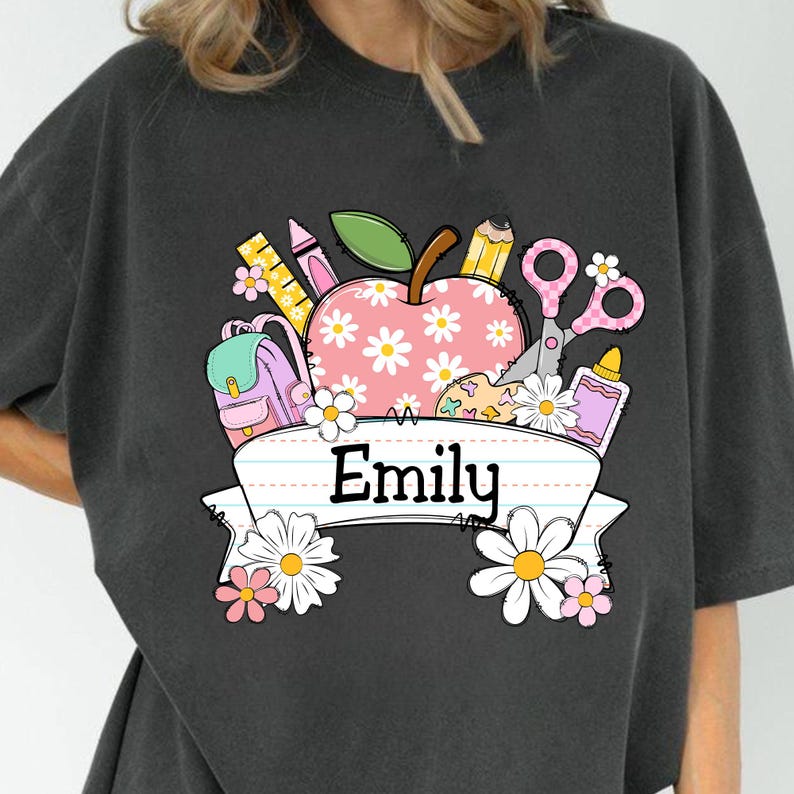 May include: Dark gray t-shirt featuring a school-themed graphic. The design includes a pink apple with daisy patterns, school supplies, and the name "Emily" on a banner. The graphic is colorful and whimsical, perfect for back-to-school.