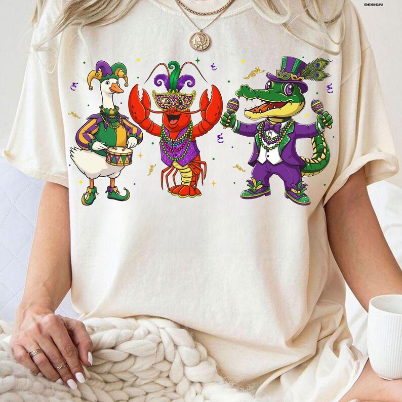 May include: Cream-colored t-shirt featuring three cartoon characters in Mardi Gras attire. A goose plays a drum, a lobster wears a mask, and an alligator holds maracas. The characters are dressed in purple, green, and gold, with decorative accents.