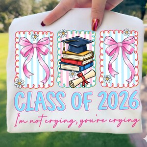 May include: White t-shirt with a graduation theme. Features three framed illustrations: two pink bows and a stack of books with a graduation cap. Text reads "CLASS OF 2026" and "I'm not crying, you're crying."