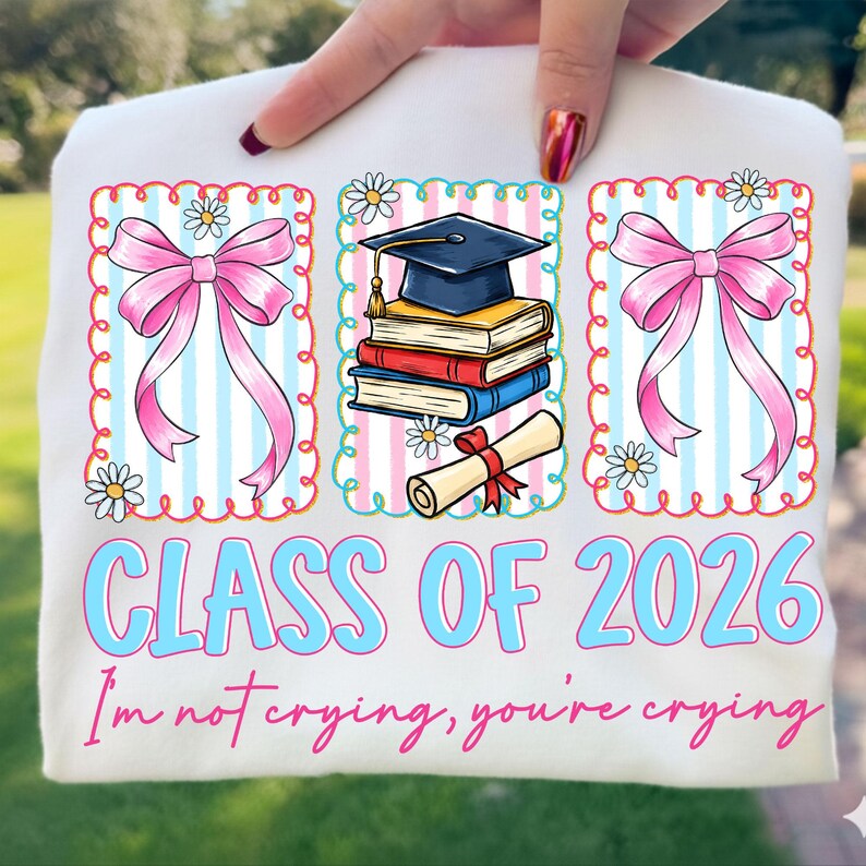 May include: White t-shirt with a graduation theme. Features three framed illustrations: two pink bows and a stack of books with a graduation cap. Text reads "CLASS OF 2026" and "I'm not crying, you're crying."