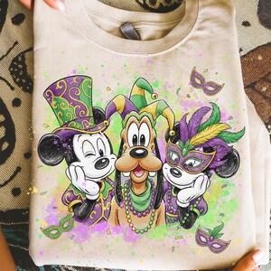 May include: Beige t-shirt featuring Mickey Mouse, Goofy, and Mickey Mouse in Mardi Gras attire. The characters wear festive hats, masks, and beaded necklaces. The design includes purple, green, and yellow accents, with a colorful splatter background.