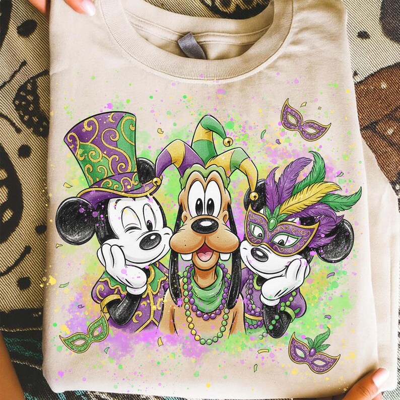 May include: Beige t-shirt featuring Mickey Mouse, Goofy, and Mickey Mouse in Mardi Gras attire. The characters wear festive hats, masks, and beaded necklaces. The design includes purple, green, and yellow accents, with a colorful splatter background.
