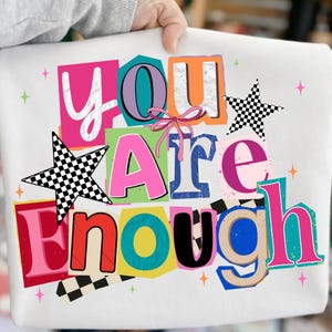 Könnte beinhalten: Weißes Sweatshirt mit dem Schriftzug "YOU ARE ENOUGH" in einem farbenfrohen Design im Collage-Stil. Das Design umfasst rosa, orange, blaue und grüne Buchstaben, karierte Sternakzente und eine rosa Schleife.