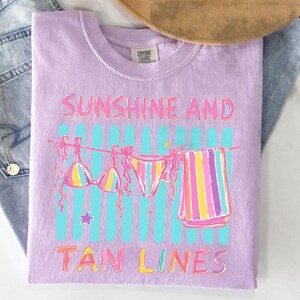 Peut inclure: T-shirt lilas Comfort Colors avec l'inscription &laquo; Sunshine and Tan Lines &raquo; en rose et jaune. Le motif repr&eacute;sente un bikini, un bas de bikini et une serviette ray&eacute;e suspendus &agrave; une corde &agrave; linge avec des rayures verticales bleues.