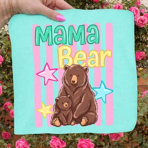 Pu&ograve; includere: Un tessuto azzurro con le parole "MAMA Bear" in verde e giallo, con un'illustrazione di un orso bruno e un cucciolo. Stelle rosa e blu e strisce rosa decorano lo sfondo.