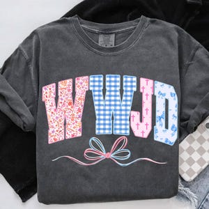 May include: Dark gray long-sleeve shirt with the letters "WWJD" in a colorful design. The "W" is floral, the second "W" is blue gingham, the "J" has pink crosses, and the "D" has blue bows. A pink and blue bow is at the bottom.