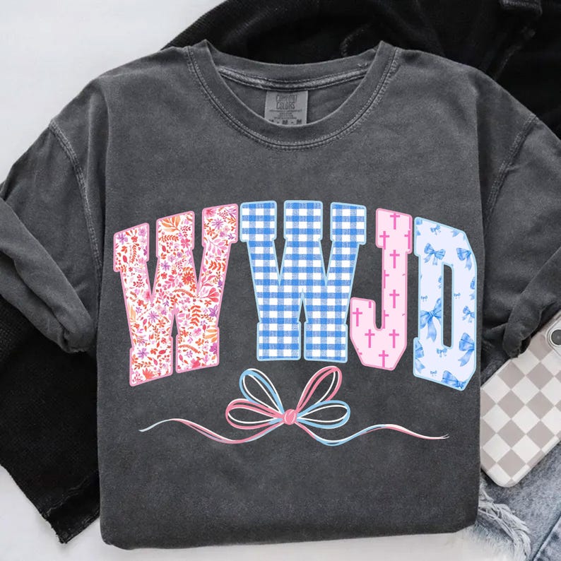 May include: Dark gray long-sleeve shirt with the letters "WWJD" in a colorful design. The "W" is floral, the second "W" is blue gingham, the "J" has pink crosses, and the "D" has blue bows. A pink and blue bow is at the bottom.