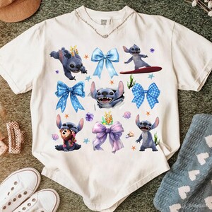 May include: White t-shirt featuring a collage of Stitch illustrations and colorful bows. The design includes Stitch in various poses, surrounded by blue, purple, and pink bows, starfish, and floral accents. The shirt has a crew neck and short sleeves.