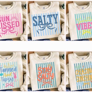 Op de afbeelding: Crèmekleurige sweatshirts met verschillende zomerse zinnen in kleurrijke tekst. Ontwerpen zijn onder meer "Sun Kissed", "Salty", "Summer Vibes", "Tanned and Tipsy", "Stay Salty" en "Chasing Sunshine", elk met gestreepte achtergronden en strikken.