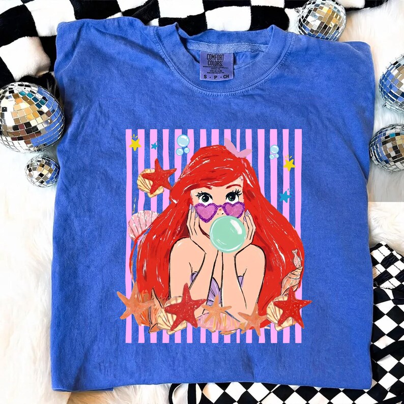 May include: A blue Comfort Colors t-shirt with a cartoon mermaid graphic. The mermaid has red hair, heart-shaped sunglasses, and a bubble. The graphic is set against pink and white stripes, with starfish and seashells. The shirt has a crew neck.