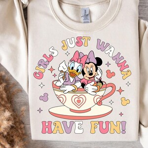 May include: A white sweatshirt with a pink, yellow, and purple graphic design. The graphic features two cartoon characters, a duck and a mouse, sitting in a teacup. The text "Girls Just Wanna Have Fun!" is written in a retro font.