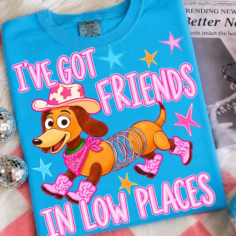 May include: A bright blue t-shirt featuring a graphic of a brown dachshund toy with a metal spring body, wearing a pink cowboy hat, a pink bandana, and pink cowboy boots. The text reads I'VE GOT FRIENDS IN LOW PLACES, surrounded by colorful stars.
