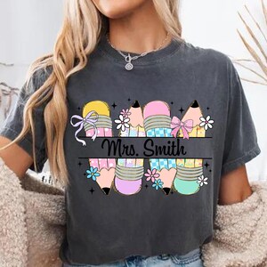 May include: Dark gray t-shirt featuring a colorful pencil design with the text "Mrs. Smith". The pencils are decorated with bows and flowers in shades of yellow, pink, blue, and purple. A cute and colorful design for teachers.