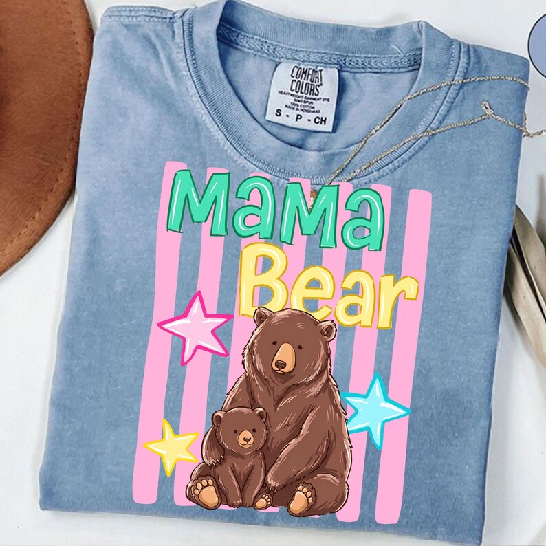 Pu&ograve; includere: Una maglietta azzurra con le parole "Mama Bear" in verde e giallo, con una grafica di un'orsa e il suo cucciolo. Strisce rosa e stelle completano il design. La maglietta &egrave; realizzata in materiale morbido.