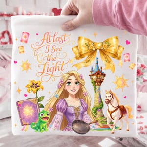 May include: White fabric featuring a colorful illustration of a princess with long blonde hair, a green chameleon, and a white horse. The text "At last I See the Light" is written in elegant script, with a golden bow and tower.
