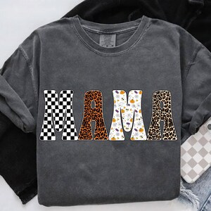May include: Dark gray sweatshirt with the word "MAMA" in large, bold letters. The letters have different patterns: checkered, leopard print, and Halloween-themed designs with ghosts, pumpkins, and bats. The sweatshirt has long sleeves.