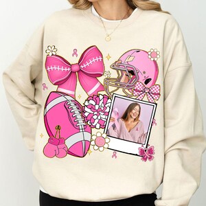 May include: Cream-colored sweatshirt featuring a pink football, helmet, bow, boxing gloves, and a photo with a woman holding a pink ribbon. The design includes pink flowers and ribbons, with a focus on breast cancer awareness.