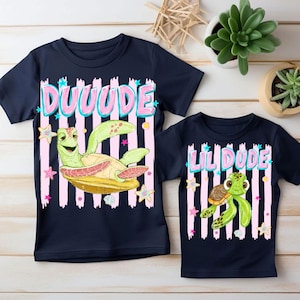 May include: Two navy blue t-shirts with turtle graphics. One shirt says "DUUUUDE" in pink letters with a green turtle graphic. The other shirt says "LIL DUDE" with a turtle graphic. Both shirts have pink and white striped backgrounds.