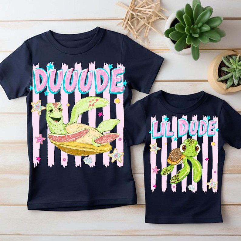 May include: Two navy blue t-shirts with turtle graphics. One shirt says "DUUUUDE" in pink letters with a green turtle graphic. The other shirt says "LIL DUDE" with a turtle graphic. Both shirts have pink and white striped backgrounds.