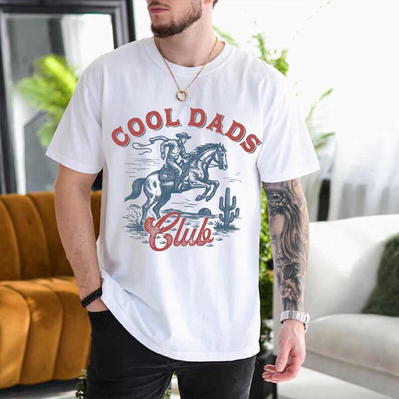 May include: White t-shirt with a graphic design featuring a cowboy riding a horse, a cactus, and the text "COOL DADS Club" in red and blue. The shirt is a casual style.