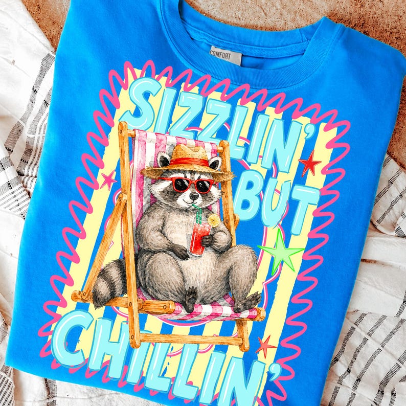 May include: Bright blue sweatshirt with a graphic of a raccoon in sunglasses and a straw hat, relaxing in a beach chair and holding a drink. The text reads "Sizzlin' But Chillin'" in a playful font, with yellow and pink accents.
