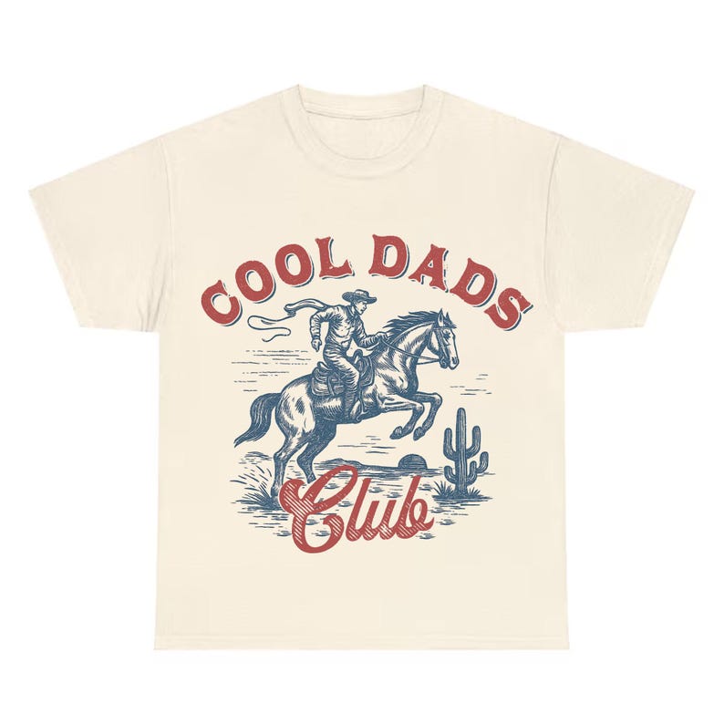 May include: Cream-colored t-shirt featuring a vintage-style graphic of a cowboy riding a horse. The design includes the text "COOL DADS Club" in red and blue, with a desert landscape backdrop.