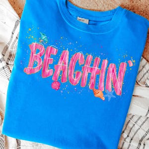 May include: A blue sweatshirt with the word "BEACHIN'" in pink, outlined in white. The design includes seashell graphics and colorful paint splatters, evoking a beach theme. The shirt is made of a soft material.