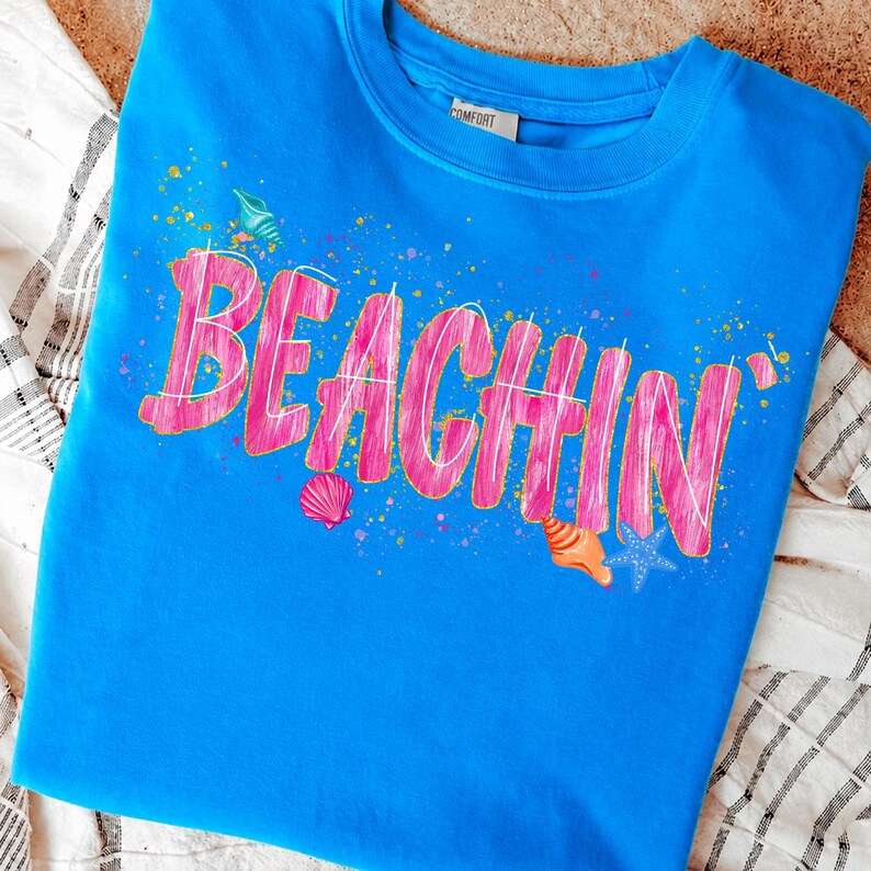 May include: A blue sweatshirt with the word "BEACHIN'" in pink, outlined in white. The design includes seashell graphics and colorful paint splatters, evoking a beach theme. The shirt is made of a soft material.