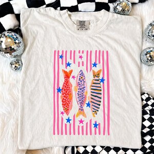 Pode incluir: Camiseta creme com um gr&aacute;fico colorido de tr&ecirc;s peixes. Os peixes s&atilde;o colocados em um fundo de listras verticais rosa e estrelas azuis. Os peixes t&ecirc;m padr&otilde;es vermelhos, azuis e amarelos.