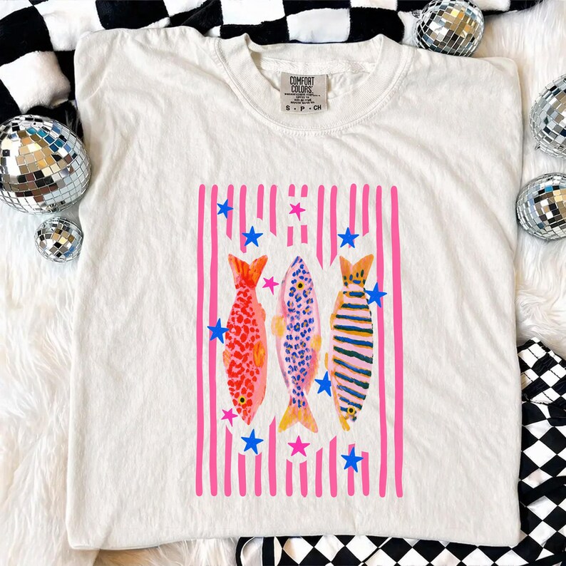 Pode incluir: Camiseta creme com um gr&aacute;fico colorido de tr&ecirc;s peixes. Os peixes s&atilde;o colocados em um fundo de listras verticais rosa e estrelas azuis. Os peixes t&ecirc;m padr&otilde;es vermelhos, azuis e amarelos.