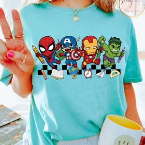 May include: A light blue t-shirt featuring a graphic of Marvel superheroes: Spider-Man, Captain America, Iron Man, and the Hulk, each holding a pencil. The design includes a black and white checkered pattern and school-themed icons.