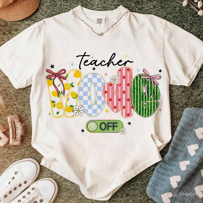 Op de afbeelding: Wit T-shirt met de tekst "Teacher Mode Off" in een speels ontwerp. De letters zijn versierd met citroenen, een blauw-wit dambordpatroon, aardbeien en watermeloen. Het shirt heeft een V-hals en een decoratieve ketting.