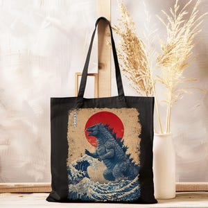 May include: Black tote bag with a blue Godzilla illustration, red sun, and ocean waves, inspired by Japanese art. The bag has long handles and a rectangular shape. The design includes Japanese text.